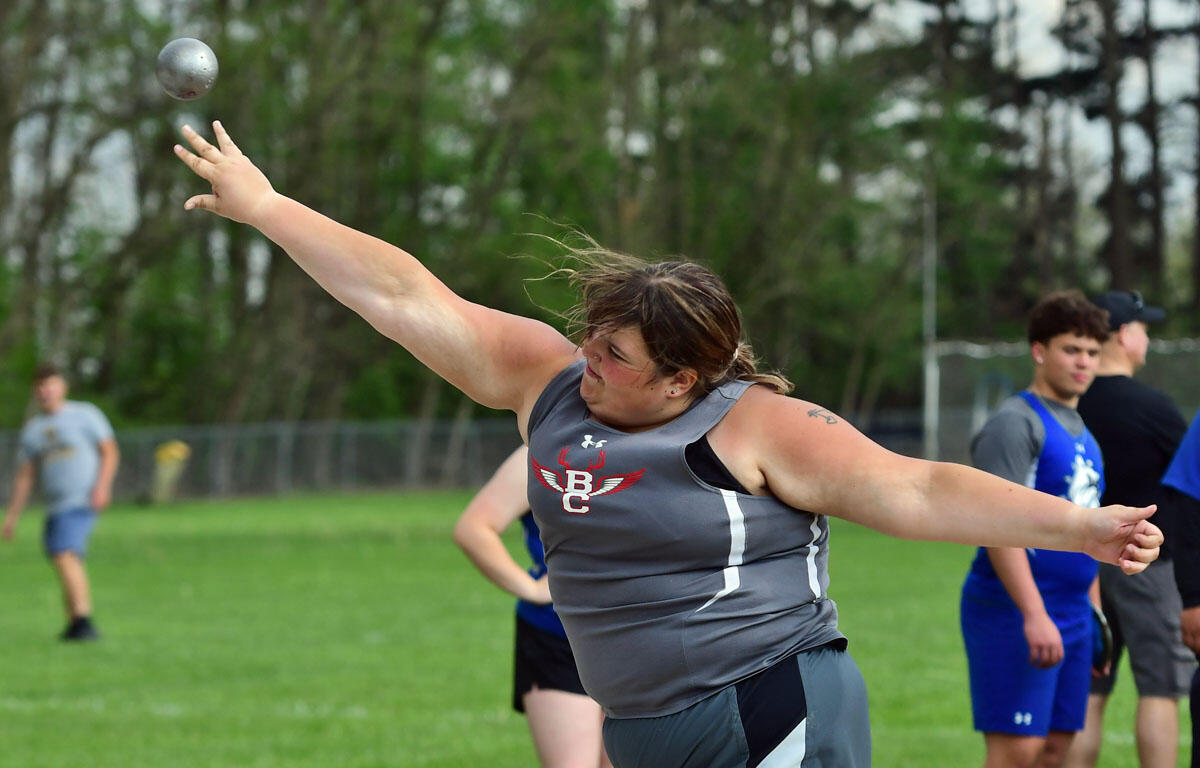 Female athlete in gray uniform throwing a shot put on a grassy field, with teammates in the background.