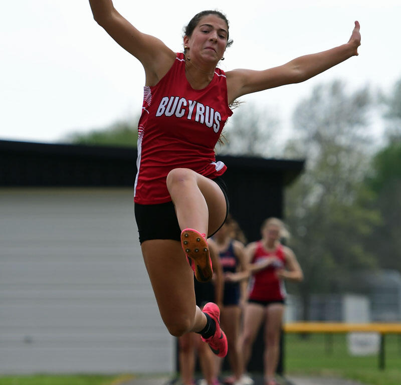 Female athlete mid-flight during a long jump, wearing a red Bucyrus jersey, on a track with onlookers in the background.