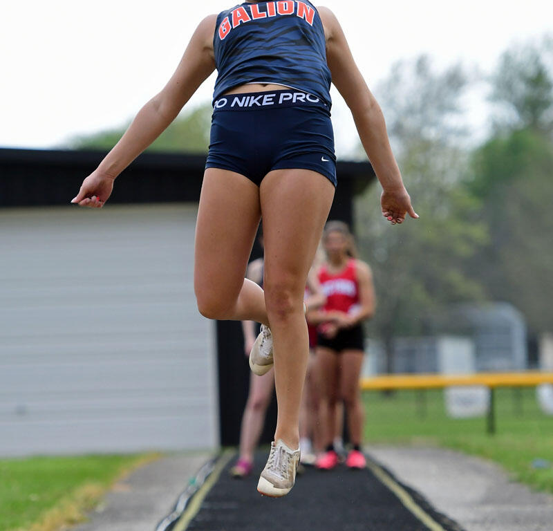Female athlete in blue uniform mid-air during a long jump toward a sand pit on an outdoor track field (competition setting).
