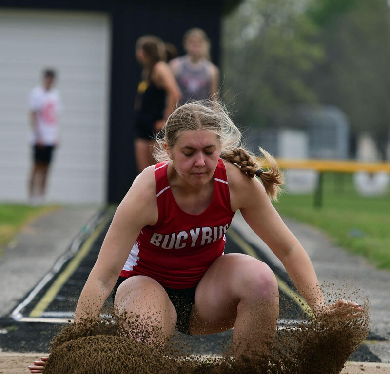 Female athlete in a red Bucyrus uniform lands in a sand pit, sand flying up during a long jump event.