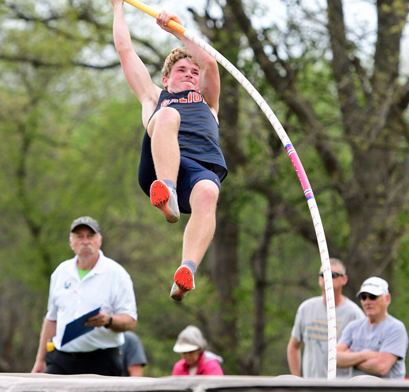 Pole vaulter mid-air clearing the bar over the padded pit during an outdoor meet, holding a yellow pole with spectators in the background.