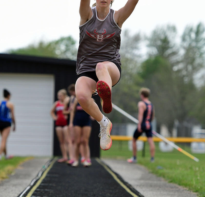 Female long jumper in a gray singlet jumping mid-air with arms raised, approaching the sand pit on a track field.