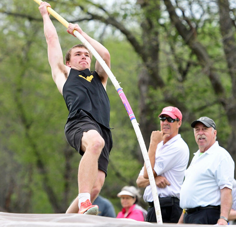 Pole vaulter in a black singlet clears the bar in an outdoor competition, with spectators watching nearby.