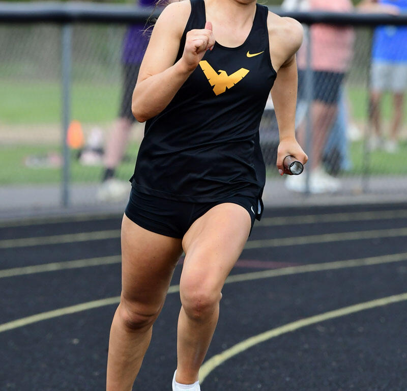 Female sprinter in a black uniform with a gold logo runs on a track, holding a baton during a relay race.