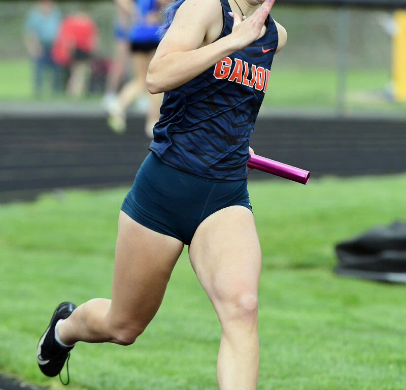 Female relay sprinter in a navy uniform runs on a track with a pink baton in hand, outdoors on a sunny day.