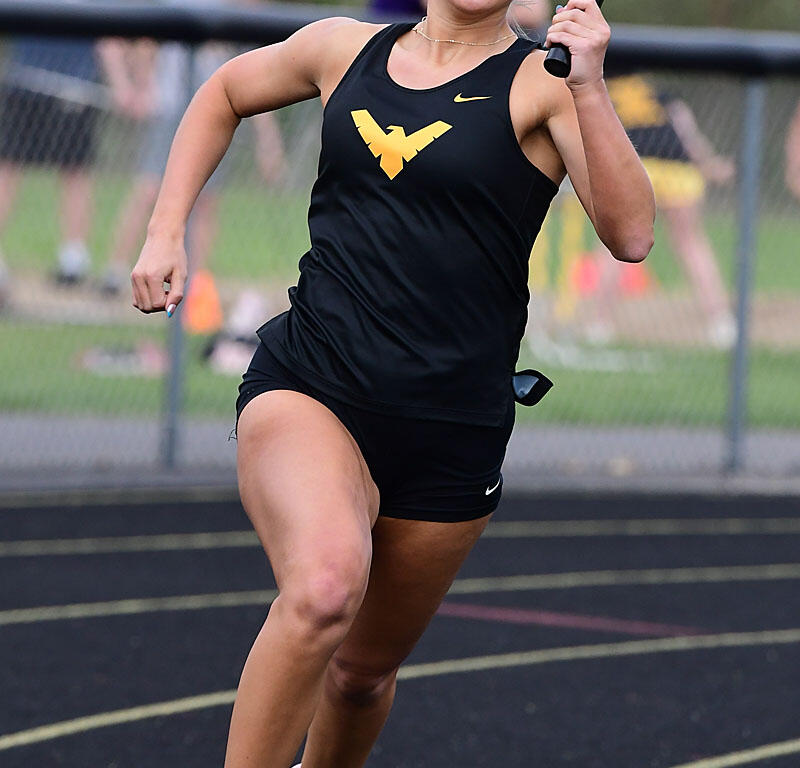 Female sprinter in a black uniform runs on a track while holding a relay baton in her right hand.