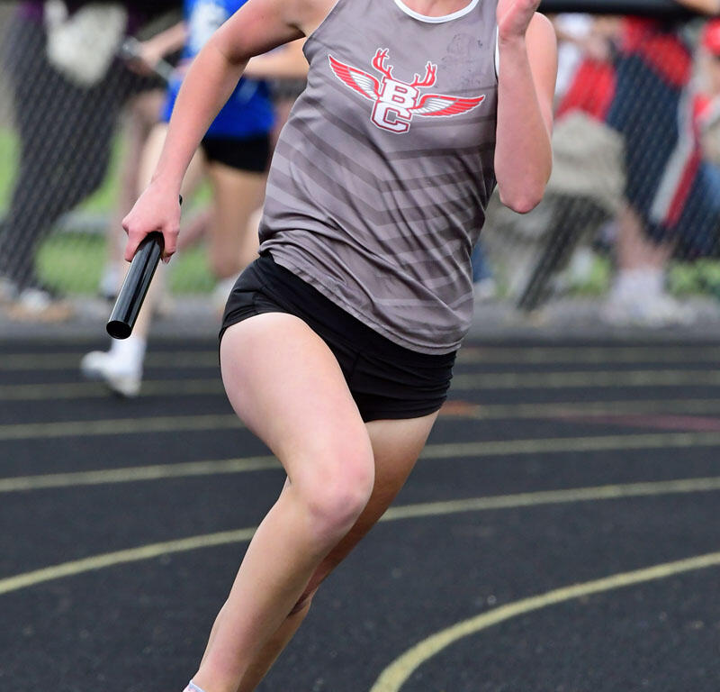 Female sprinter in a gray relay jersey runs on a track, holding a baton with spectators blurred in the background.