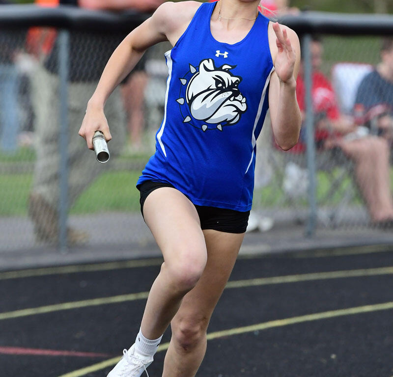 Young female baton runner in a blue jersey racing on a track, reaching for the finish line with a baton in hand and crowd in background.