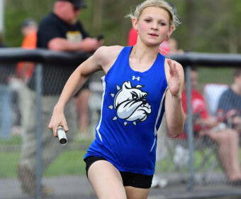 Young female baton runner in a blue jersey racing on a track, reaching for the finish line with a baton in hand and crowd in background.