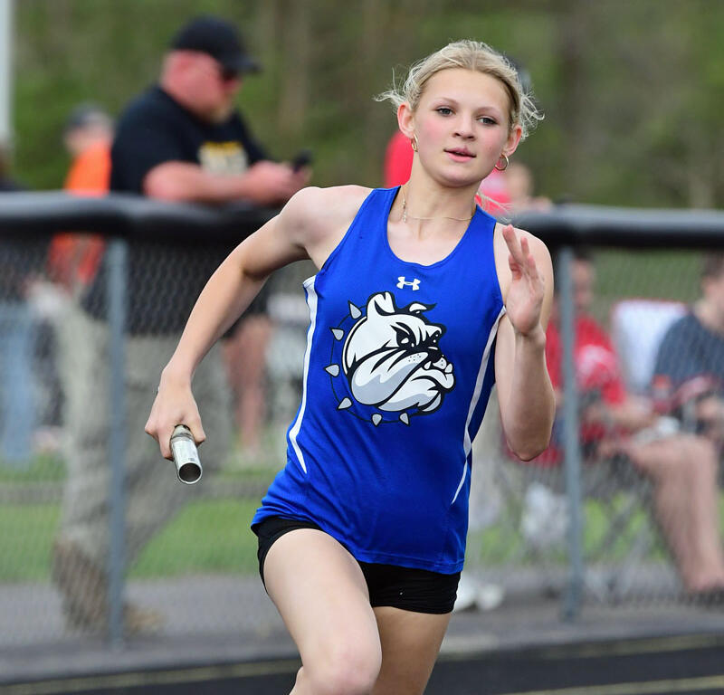 Young female baton runner in a blue jersey racing on a track, reaching for the finish line with a baton in hand and crowd in background.