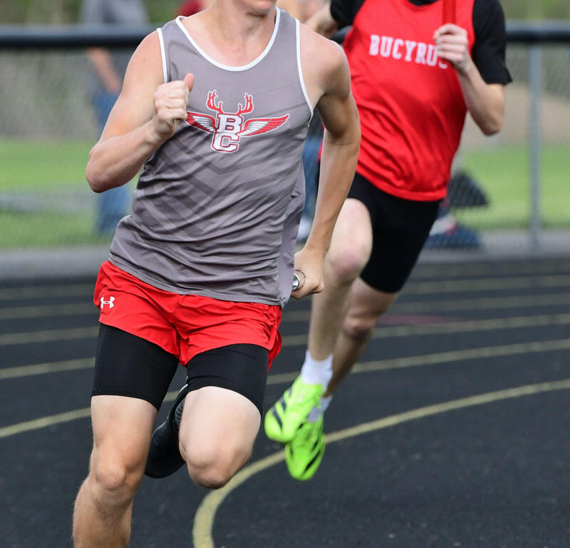 Two male runners sprinting on a curved outdoor track; foreground runner wears a gray sleeveless top and red shorts, chasing the finish line while another runner in a red Bucyrus jersey follows behind.
