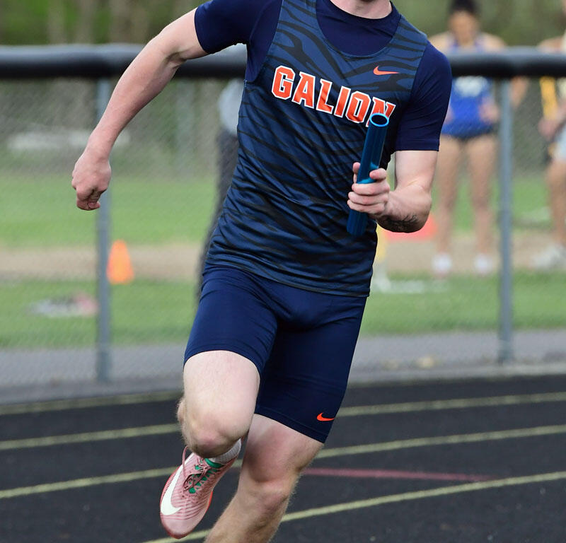 Male sprinter in a blue 'GALION' jersey runs on a track, holding a relay baton during a race.