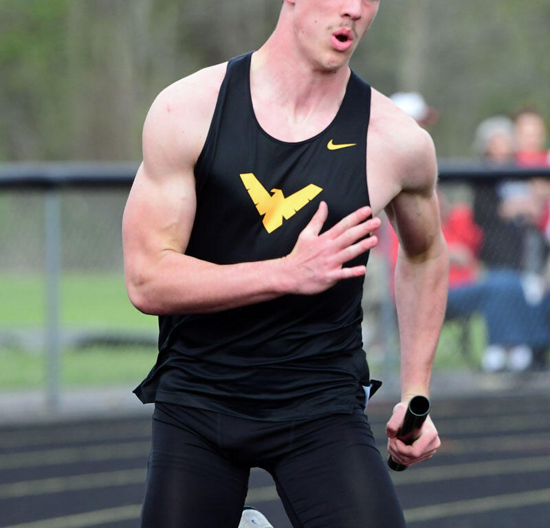 Male sprinter in a black singlet with a gold logo, running on a track while holding a relay baton.
