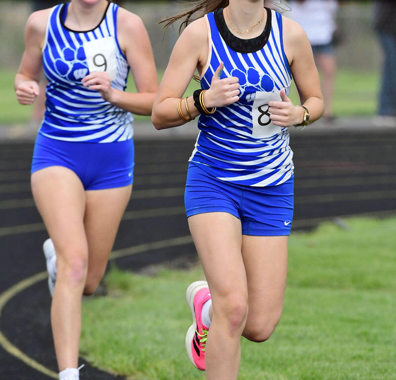 Two young female runners in blue and white striped uniforms sprint on a track, bib numbers 8 and 9 visible.