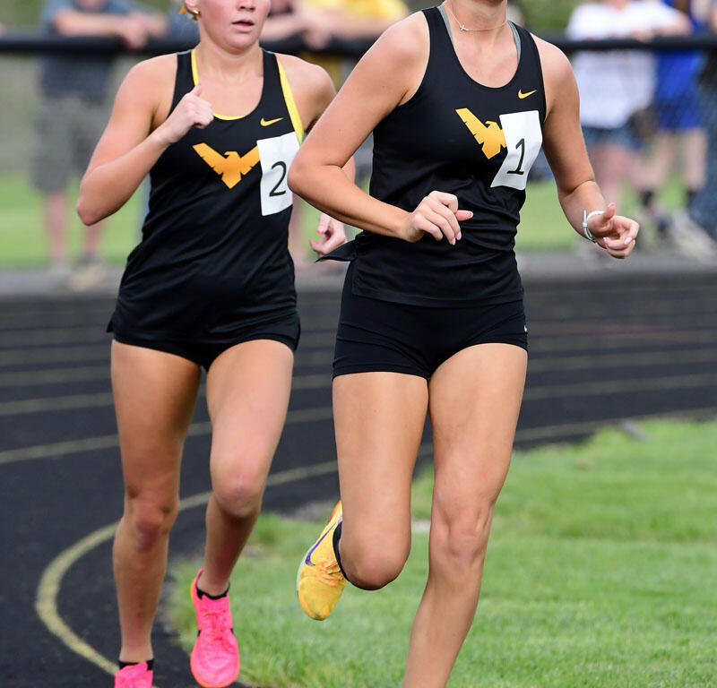 Two female sprinters in black uniforms with yellow logos race on an outdoor track, bib numbers 1 and 2 visible in front of a blurred crowd.