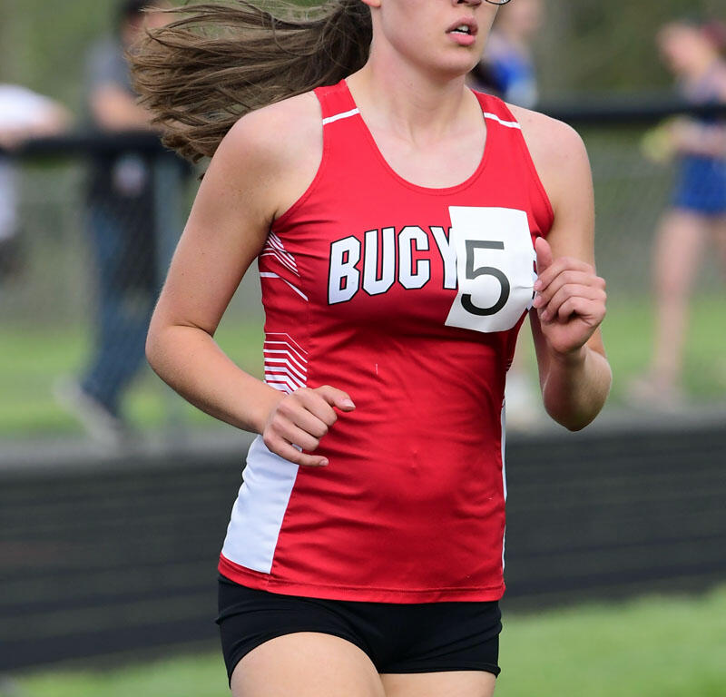 Female runner in a red athletic top with bib number 5, mid-race on a grassy track field with glasses on.