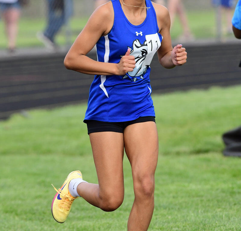 Young female athlete sprinting on a track in a blue uniform, braided hair with blue ribbons, wearing yellow sneakers with white socks.