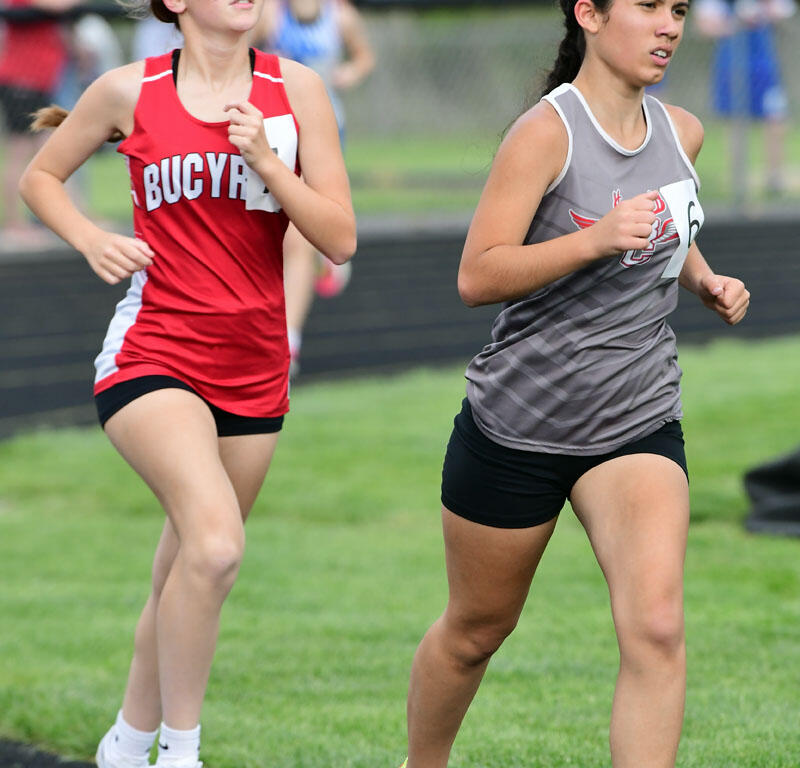 Two female runners in a track race, one in a red uniform with sunglasses and the other in gray, on a track field.