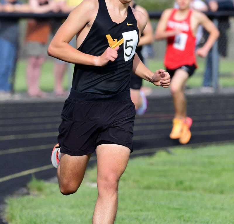 Male sprinter in a black sleeveless top and shorts, number 5, running on an outdoor track with spectators in the background.
