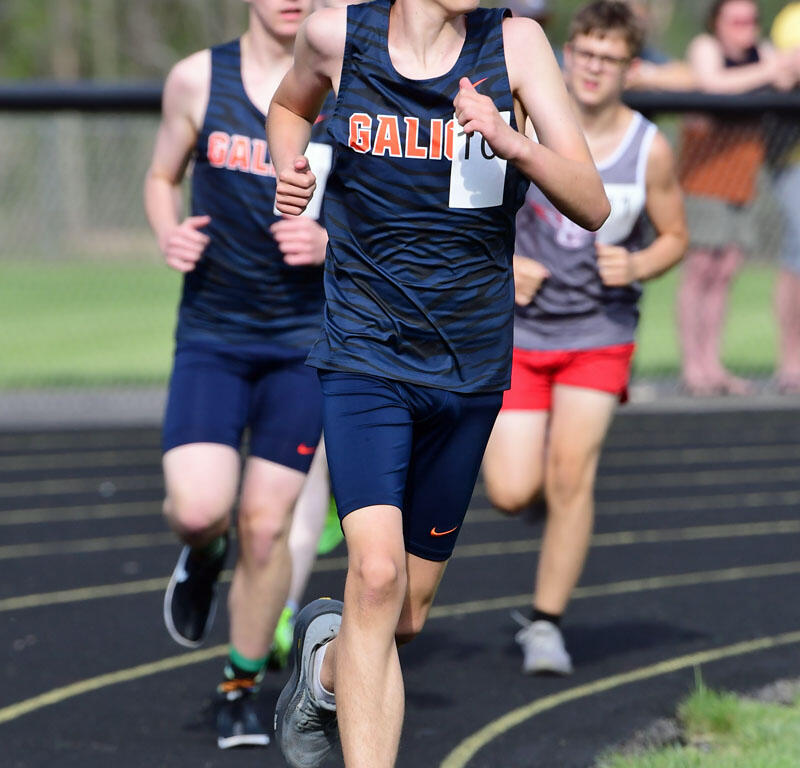 Young male runner in a navy blue singlet leads a track race on an outdoor track, with teammates behind him and spectators along the fence.