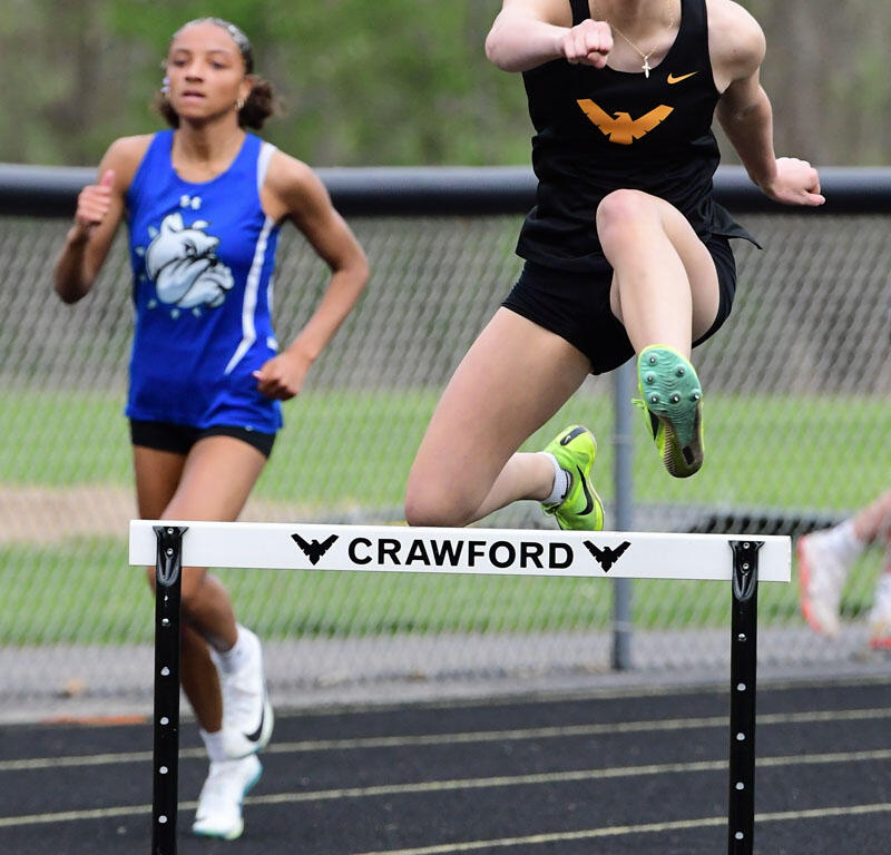 Two female runners in a hurdle race; the athlete in front is mid-jump over a Crawford hurdle wearing a black kit, green shoes, and a cross necklace, as another runner in blue follows behind on a track.