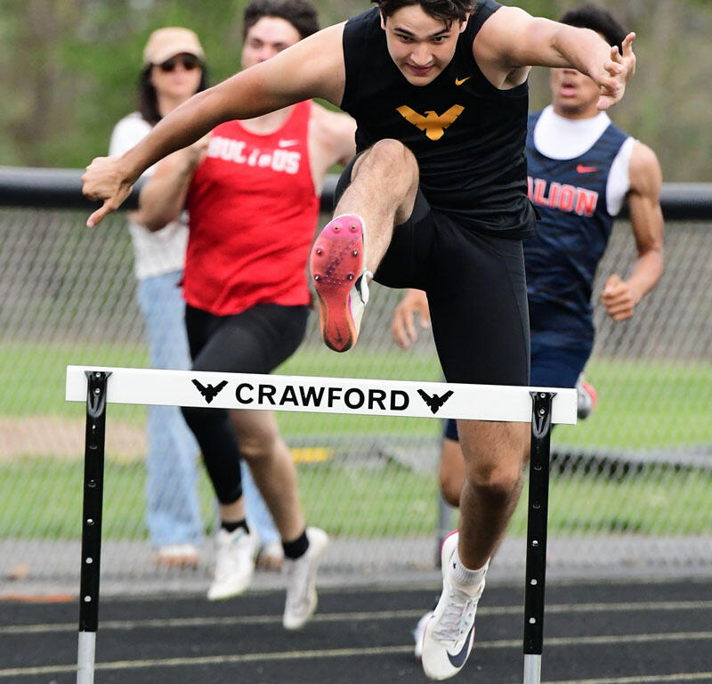 Male sprinter clears a hurdle on a track during a race, mid-air with teammates behind him and 'CRAWFORD' on the hurdle rail