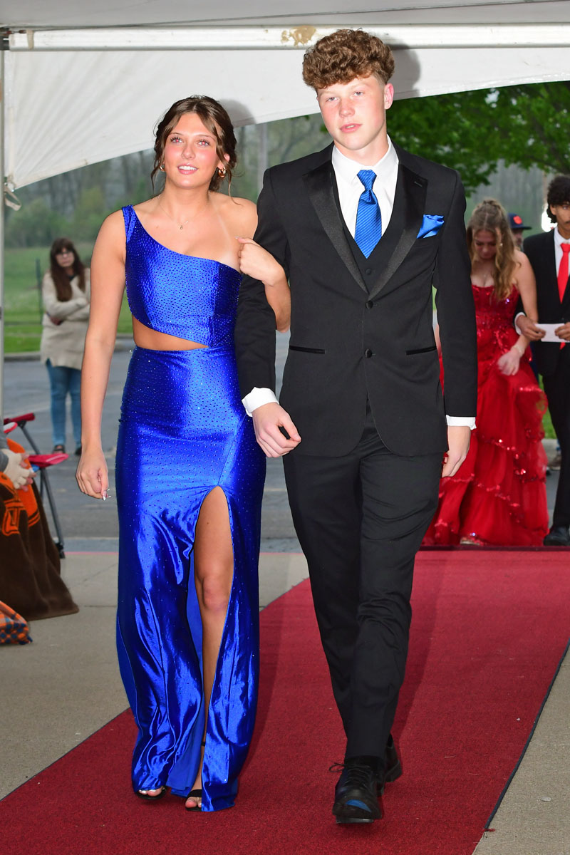 Teen couple walking arm-in-arm on a red carpet at a formal event; girl in a royal blue gown with a slit, boy in a black suit with a blue tie.