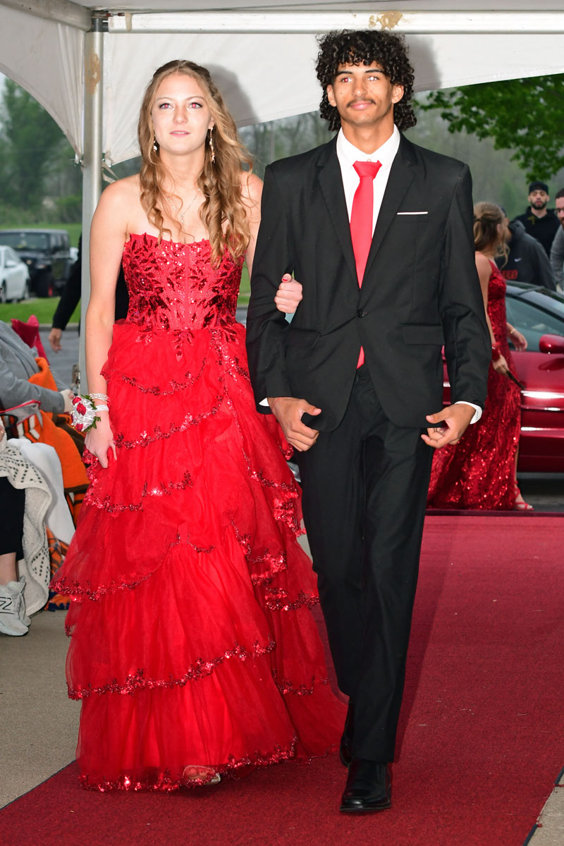 Couple walking down a red carpet at a formal event: woman in a red sequined gown and man in a black suit with a red tie under a white canopy.