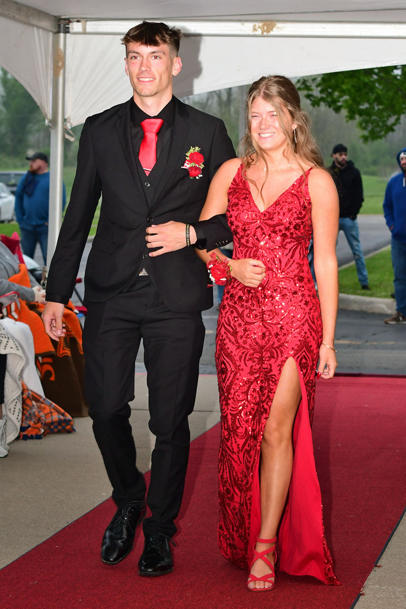 Young couple walking arm-in-arm on a red carpet; man in a black suit with red tie and boutonniere, woman in a red sequined gown with slit.