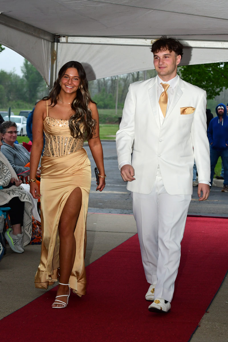 Young couple walking a red carpet at an indoor event; woman in a gold sequined gown with a high slit, man in a white suit with gold tie.