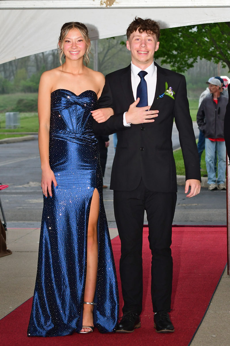 Prom-ready couple posing on a red carpet: girl in a blue sequined strapless gown with a slit, and a boy in a black suit with a blue tie beside her.