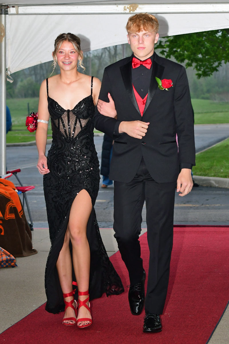 Couple walking arm-in-arm on a red carpet; girl in a black sequined gown with a high slit and red heels, boy in a black tuxedo with red bow tie and boutonniere under a white tent.