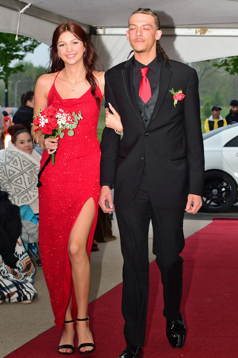 Couple walking a red carpet: woman in a red glitter dress holding a bouquet, man in a black suit with a red tie.