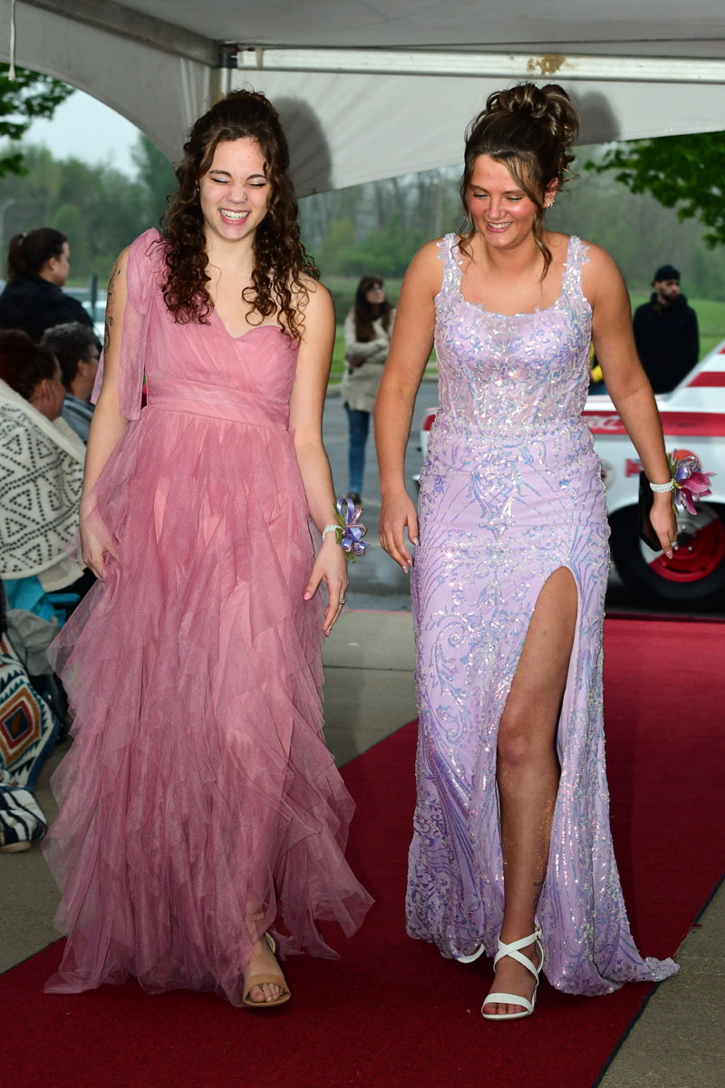 Two young women in pink and lilac formal gowns walk along a red carpet, smiling and posing under a canopy.