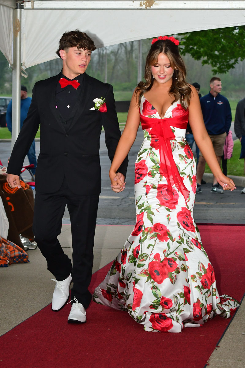 Couple walking hand in hand on a red carpet; the man in a black suit with a red bow tie and boutonniere, the woman in a white floral gown with a red sash and flower headband under a white canopy.