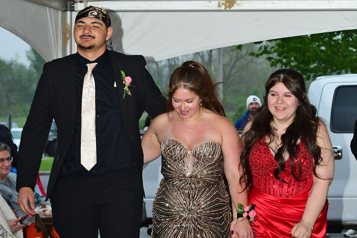 Three young people in formal attire walk arm-in-arm at a school prom or formal event, smiling as they pose together under a canopy.