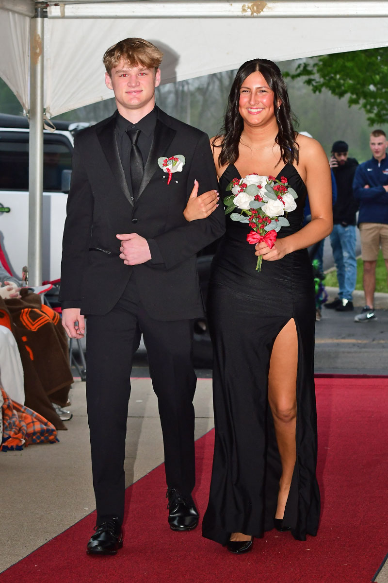Teen couple walking the red carpet at prom: the boy in a black suit and the girl in a black gown holding a bouquet, smiling.