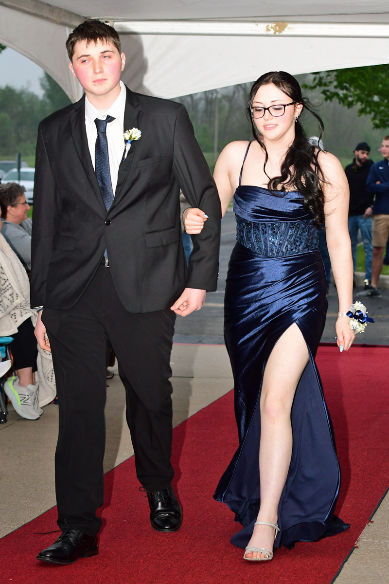 Young couple walking arm-in-arm down a red carpet; man in a black suit with boutonniere, woman in a navy blue evening gown with a high slit.