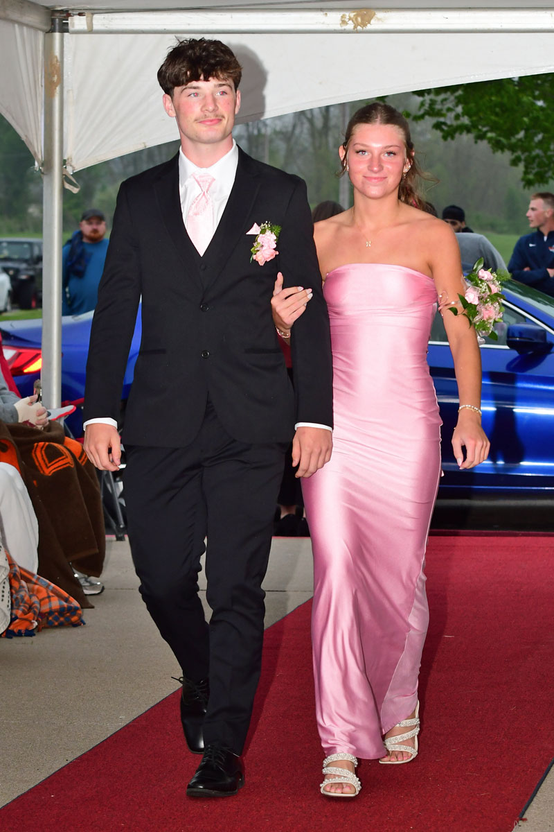 Couple walking arm-in-arm down a red carpet at a formal event; man in a black suit with a pink tie, boutonniere, and the woman in a pink strapless gown with a corsage on her wrist.