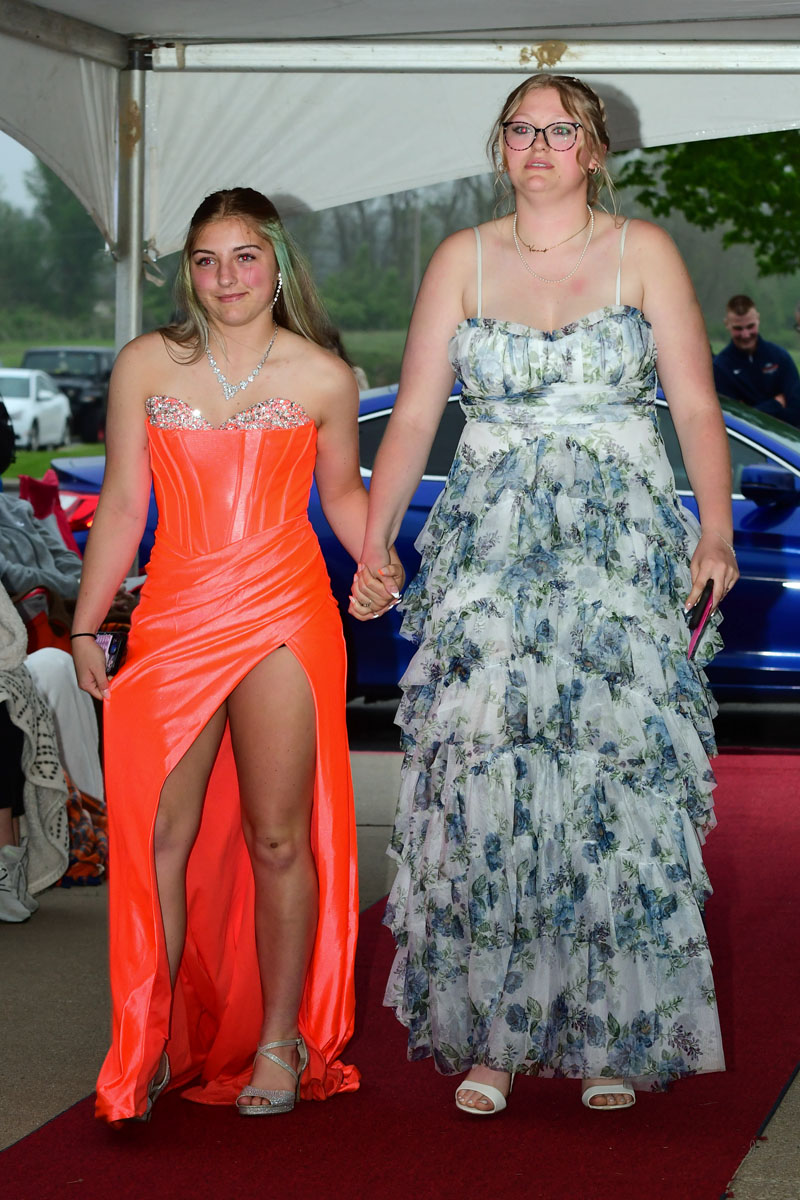 Two young women in formal gowns walk hand in hand down a red carpet under a white canopy, one in a bright orange slit dress and the other in a blue floral ball gown.