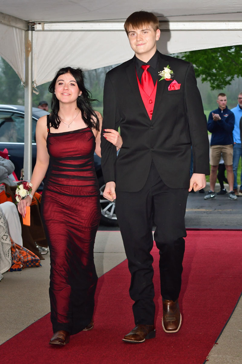 Young couple walking arm in arm on a red carpet at a formal event; girl wears a burgundy gown with a corsage, boy wears a black suit with a red tie.