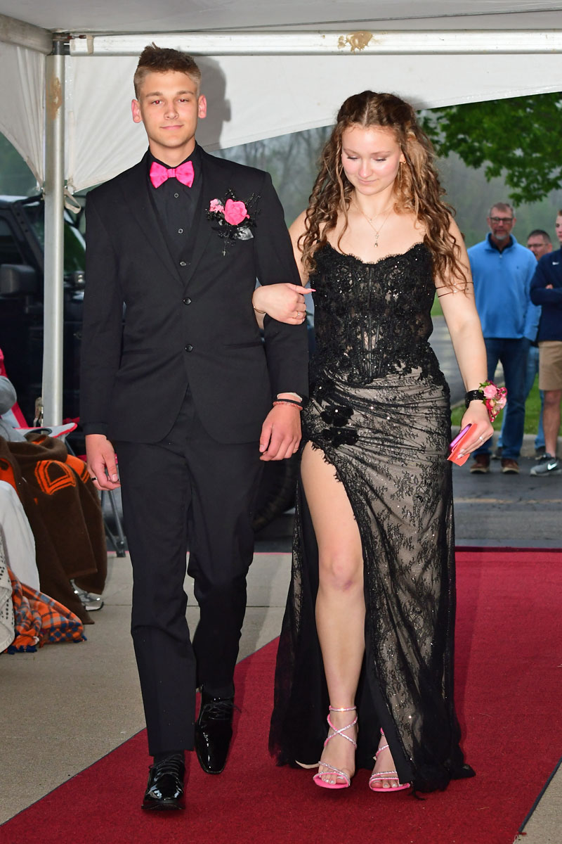 Young couple walking arm-in-arm on a red carpet at a formal event; the boy wears a black tux with a pink bow tie and boutonniere, and the girl wears a black lace gown with a high slit.
