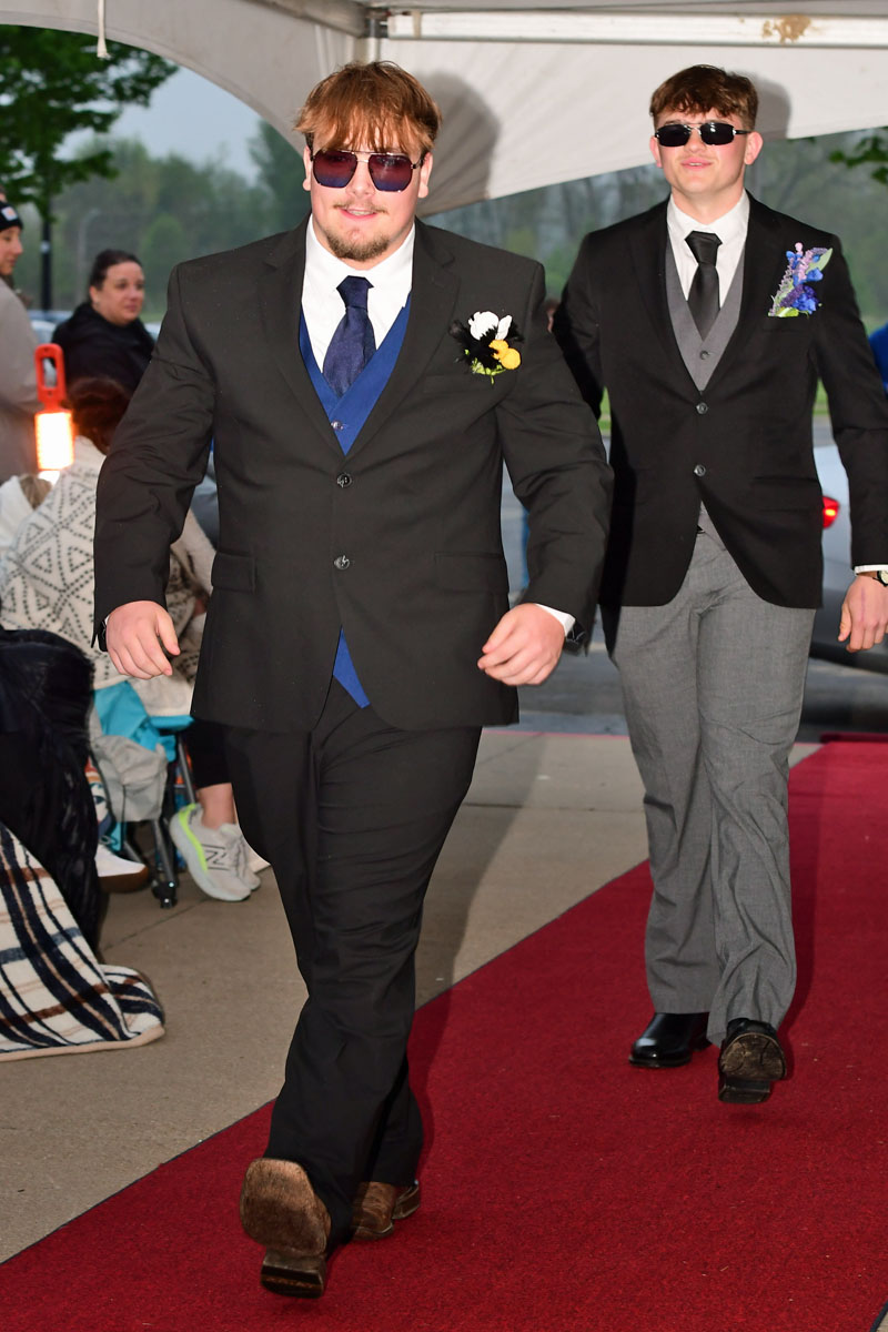 Two men in formal suits walk the red carpet under a tent; the front man wears a black suit with a blue vest, sunglasses, and a boutonnière, while the man behind wears gray pants and a black tie.