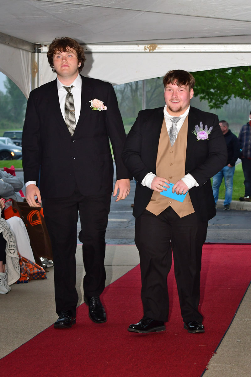 Two men in formal suits walk down a red carpet under a white tent at an outdoor event; one wears a pink boutonniere.