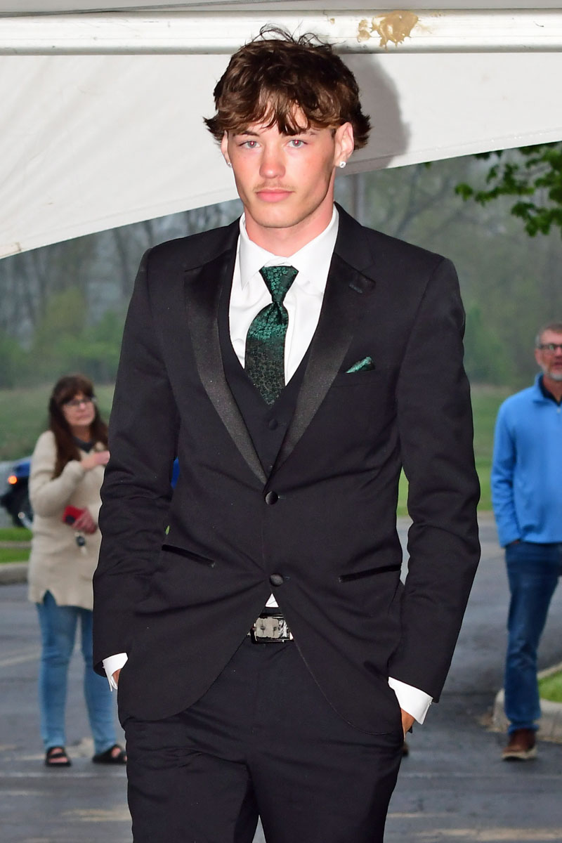 Young man in a black suit with a green tie and white shirt, hands in pockets, outdoors.