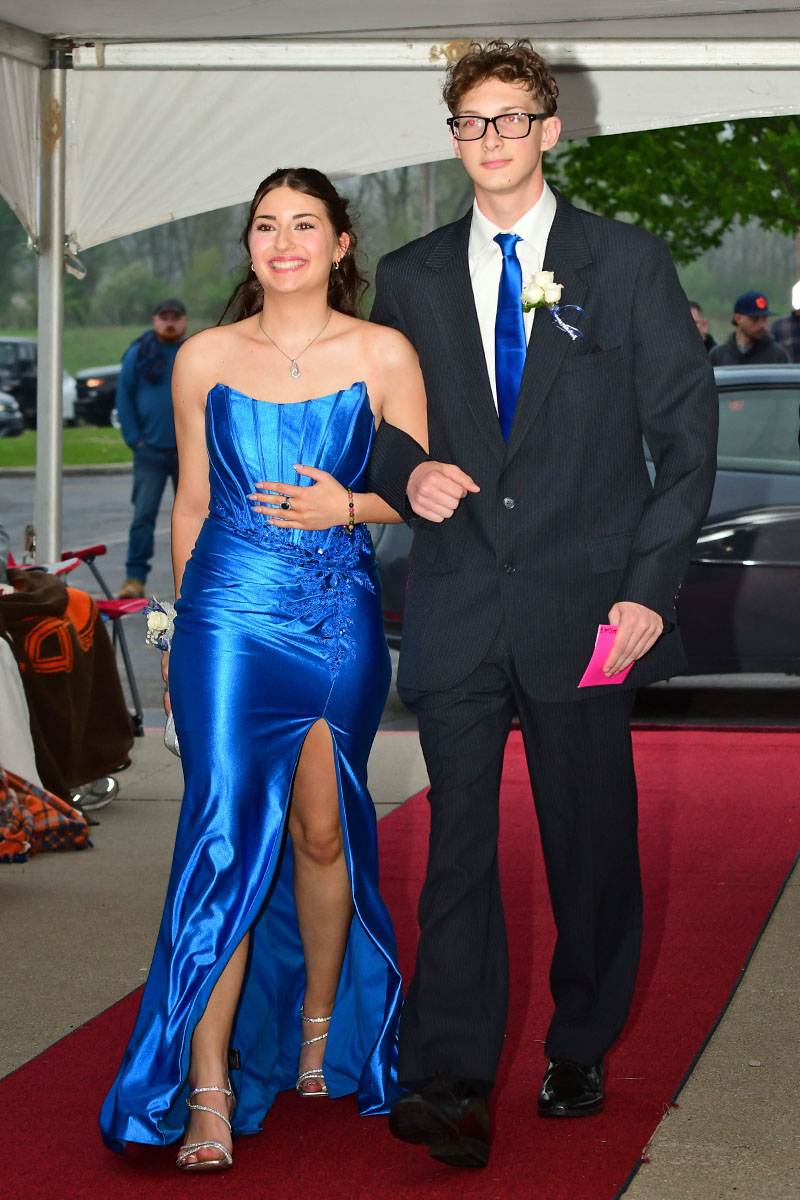 Young couple walking on a red carpet; girl in a blue strapless gown, guy in a dark suit with blue tie escorting her at prom.