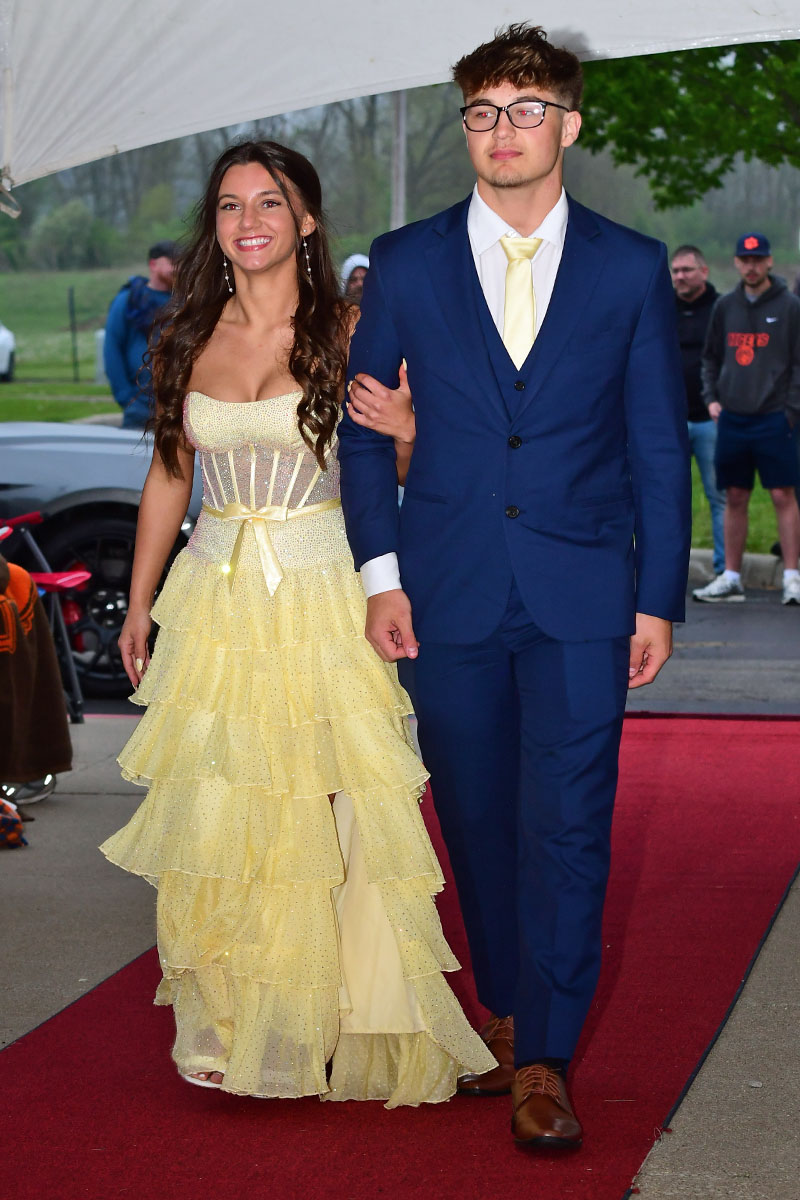 Young couple walking arm-in-arm down a red carpet; woman in a yellow tiered gown, man in a blue suit with a pale yellow tie, outdoors under a tent.