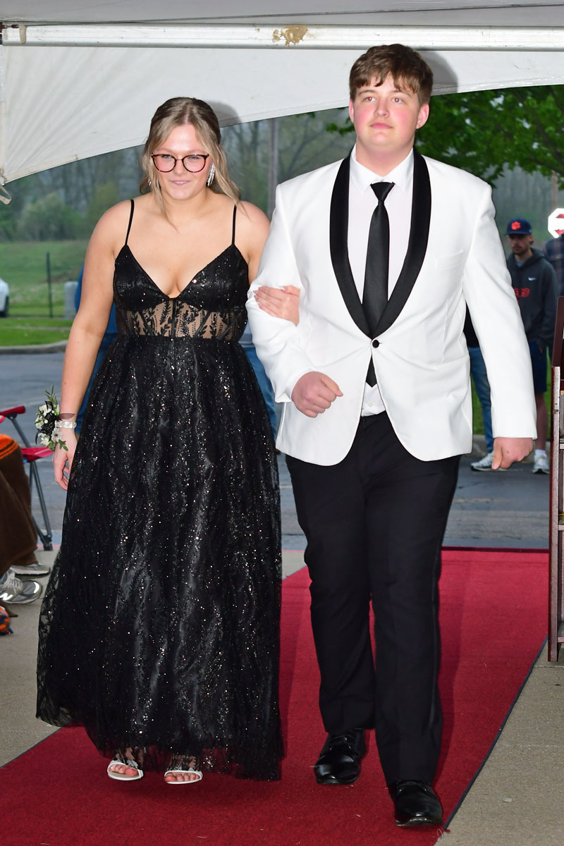 Couple walking arm-in-arm on a red carpet; girl in a glittery black gown and glasses, boy in a white tuxedo jacket and black tie.