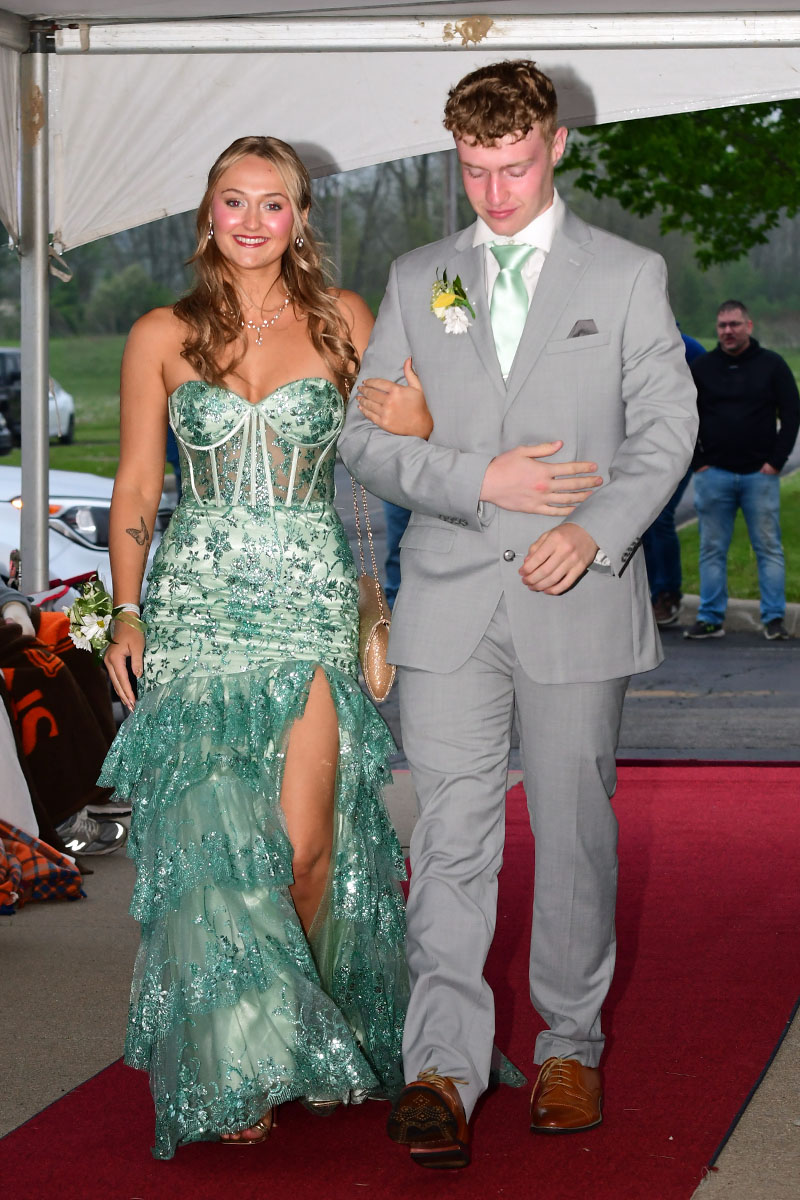 Couple walking arm-in-arm on a red carpet; woman in a teal sequined gown, man in a light gray suit with a boutonniere under a tent.