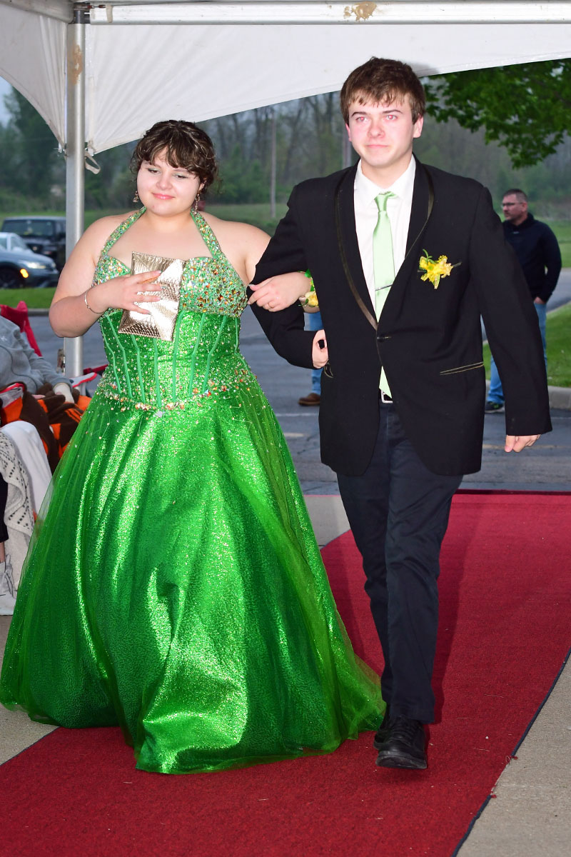 A young couple walk arm-in-arm on a red carpet: girl in a glittering green ball gown and clutch, boy in a black suit with a lime green tie.